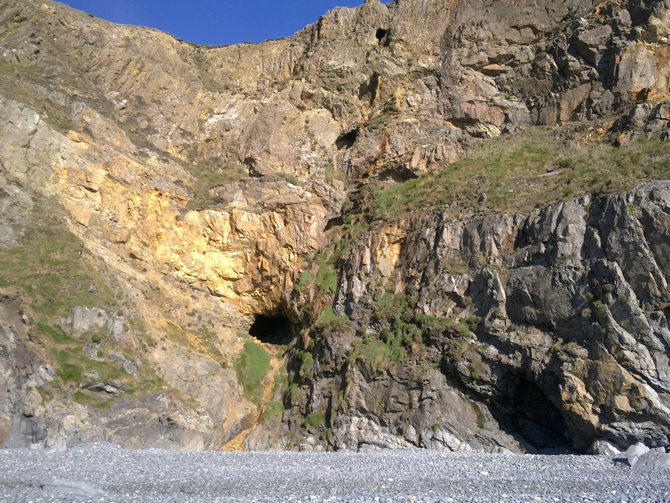 Cave-like mine adit openings in a weathered coastal rock face above a pebble beach