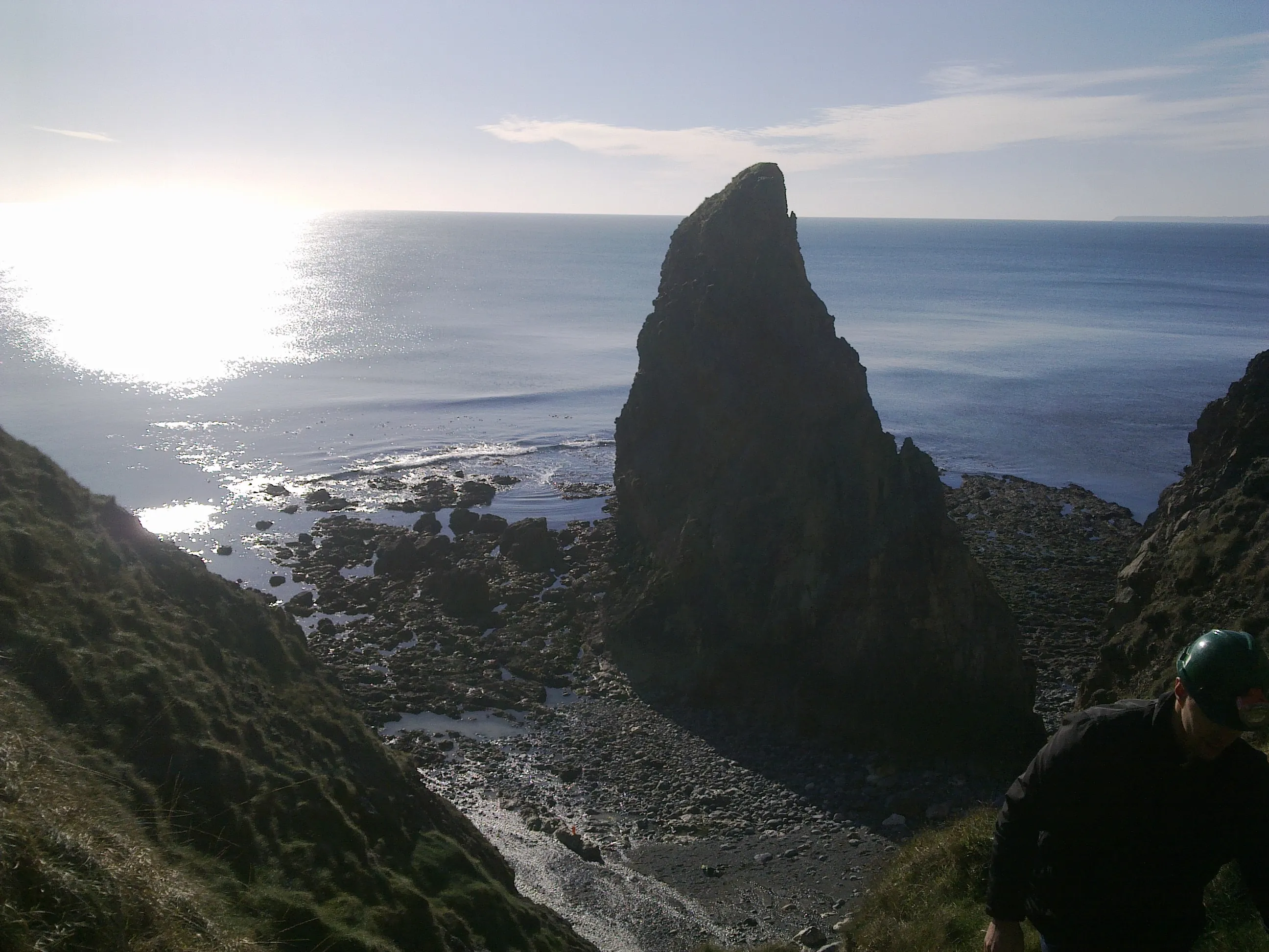 Helmeted surveyor overlooks a rocky sea stack and boulder beach along a coastal cliff
