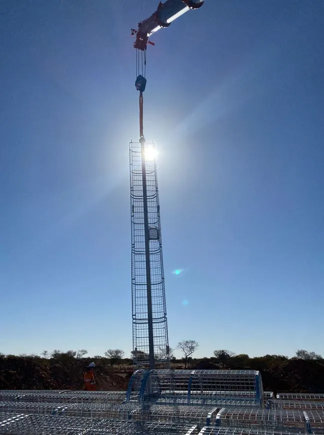 Crane lifting a tall caged ladderway section into place at a mine site under a clear sky