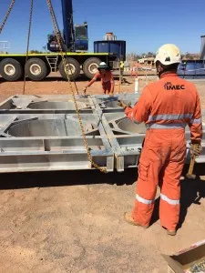 Workers in orange overalls guide a mobile crane lowering a steel fan housing onto a trailer at a mine site