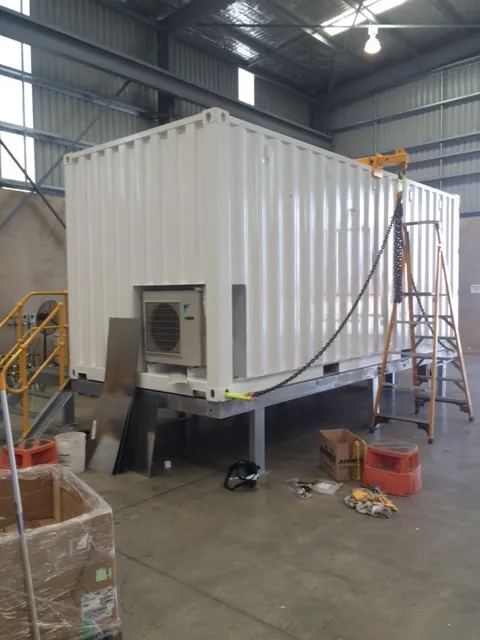 White modular container unit with air conditioning, mounted on a steel skid frame in a workshop