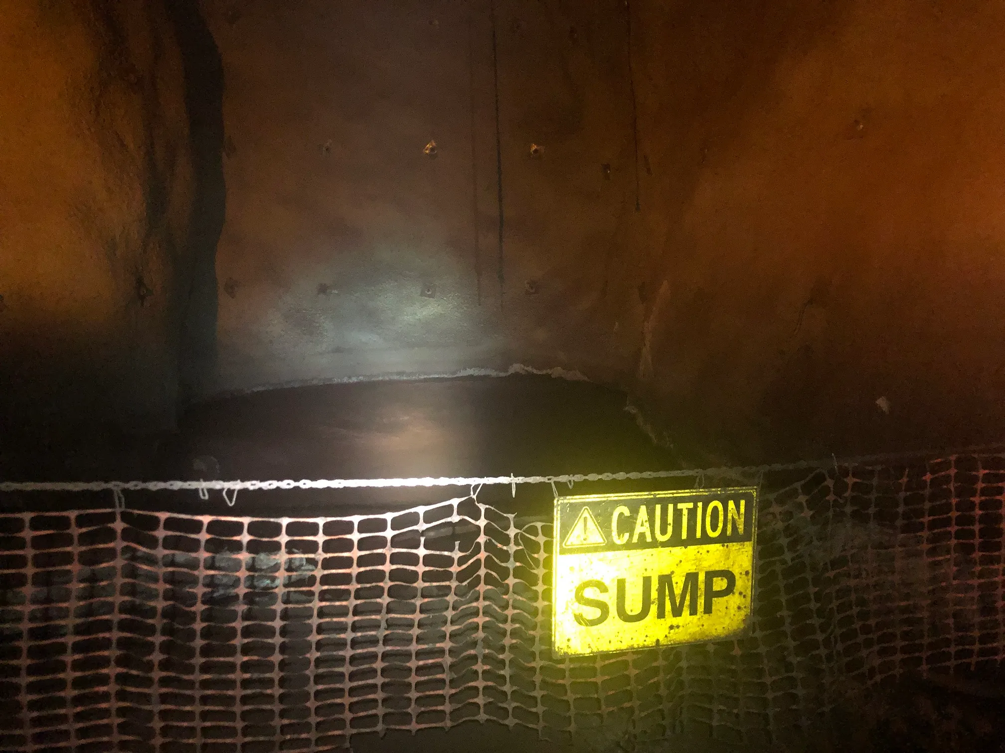 Underground mine tunnel with a barrier net and yellow “Caution Sump” sign near a dark pool