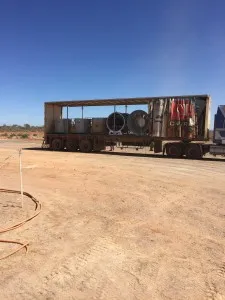 Flatbed trailer carrying large cylindrical vent fan components on a dusty mine site