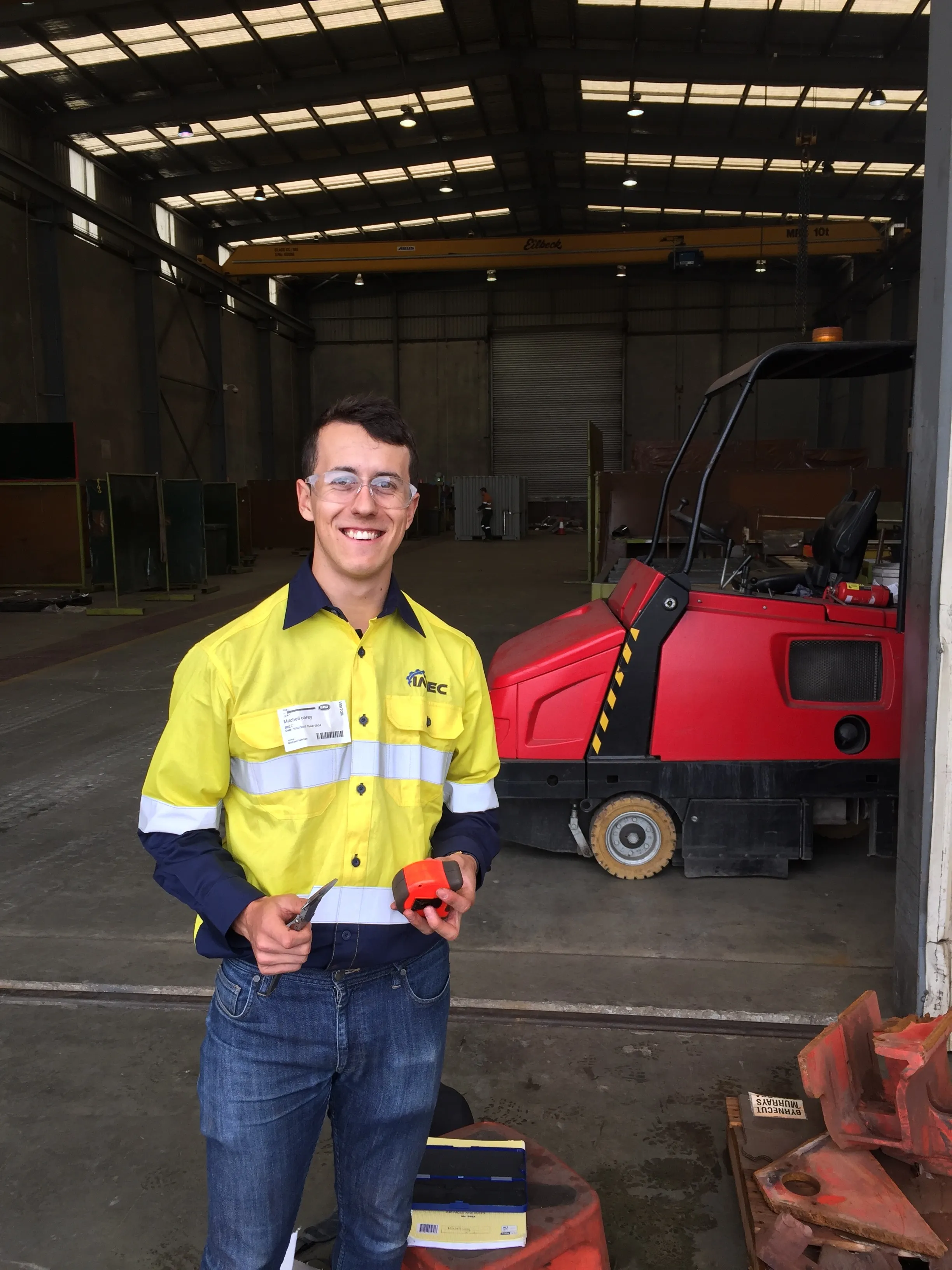 Man in hi-vis shirt and safety glasses holding a gas detector beside a red ride-on floor sweeper in a workshop