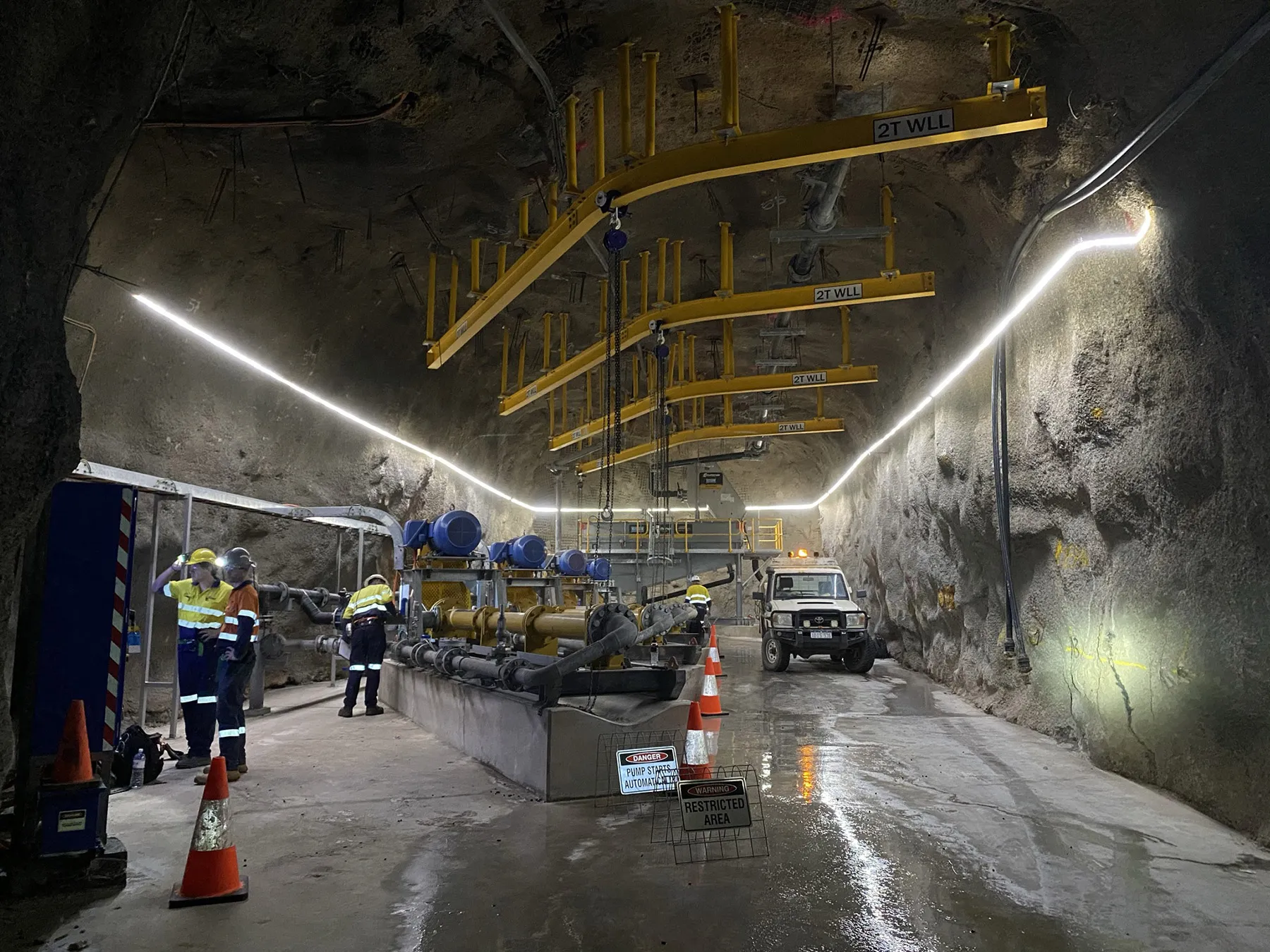 Underground mine pump station with overhead gantry cranes, workers and a utility vehicle in the tunnel