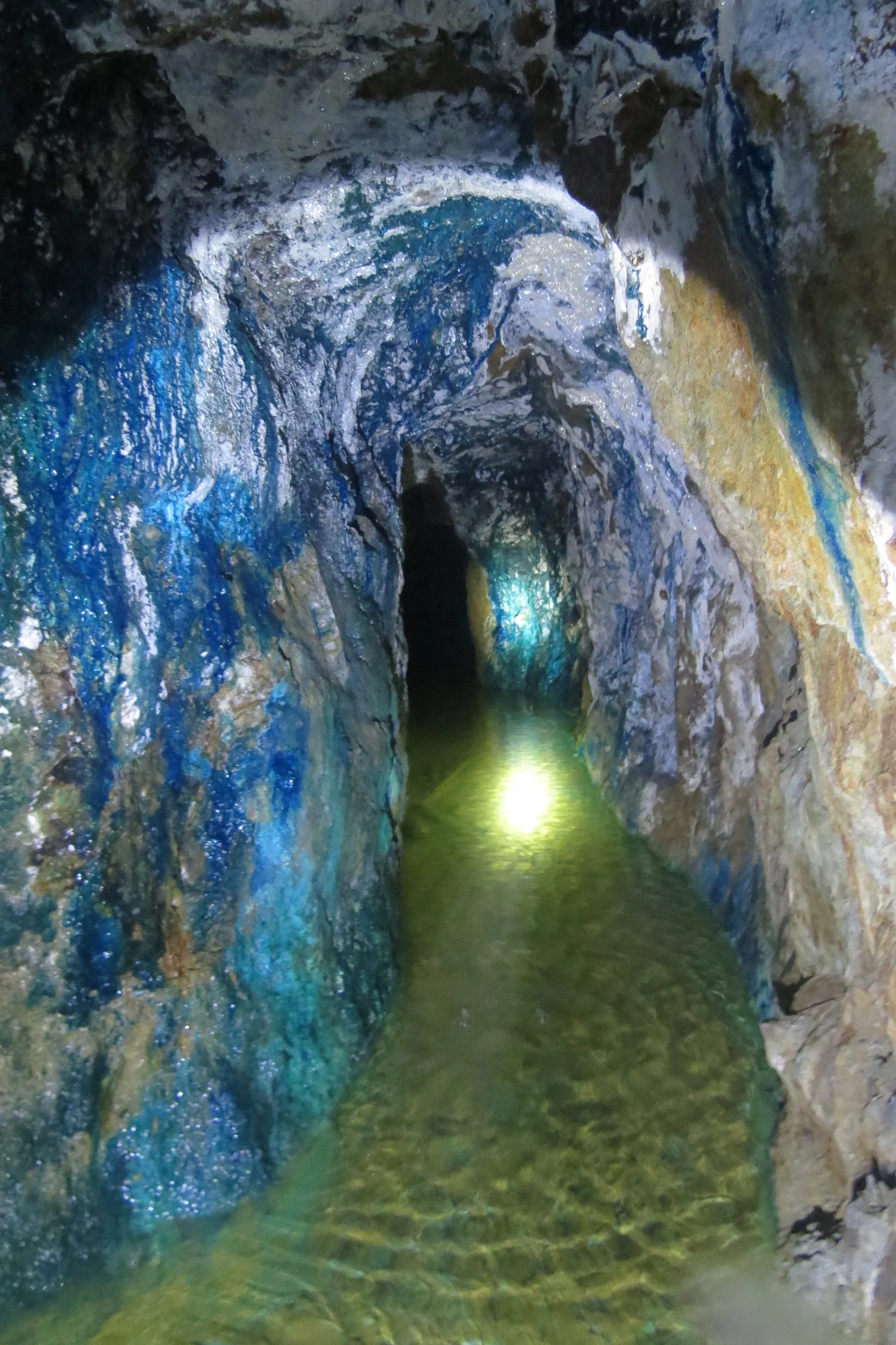 Flooded underground mine tunnel with copper mineral staining and headlamp reflecting on green water