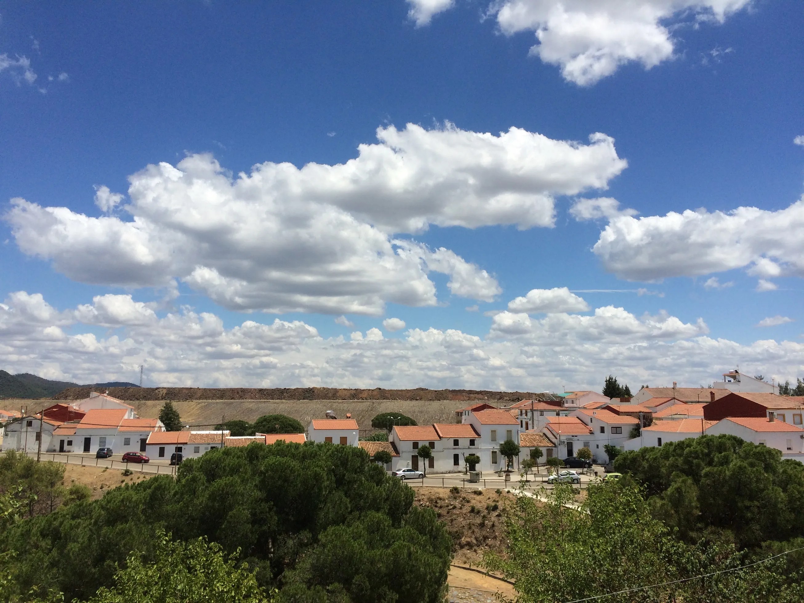 White-roofed village with terracotta-tiled houses beside a mine waste embankment under a cloud-filled sky