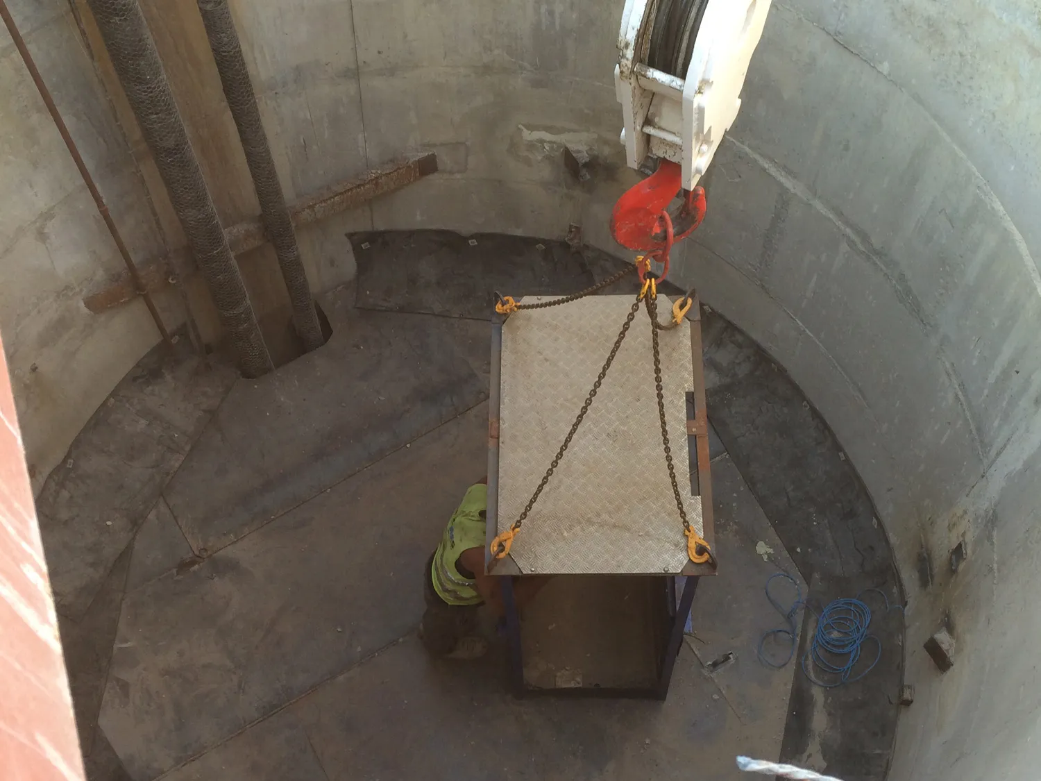 Crane lowering a chained steel platform into a concrete mine shaft as a worker guides it into position
