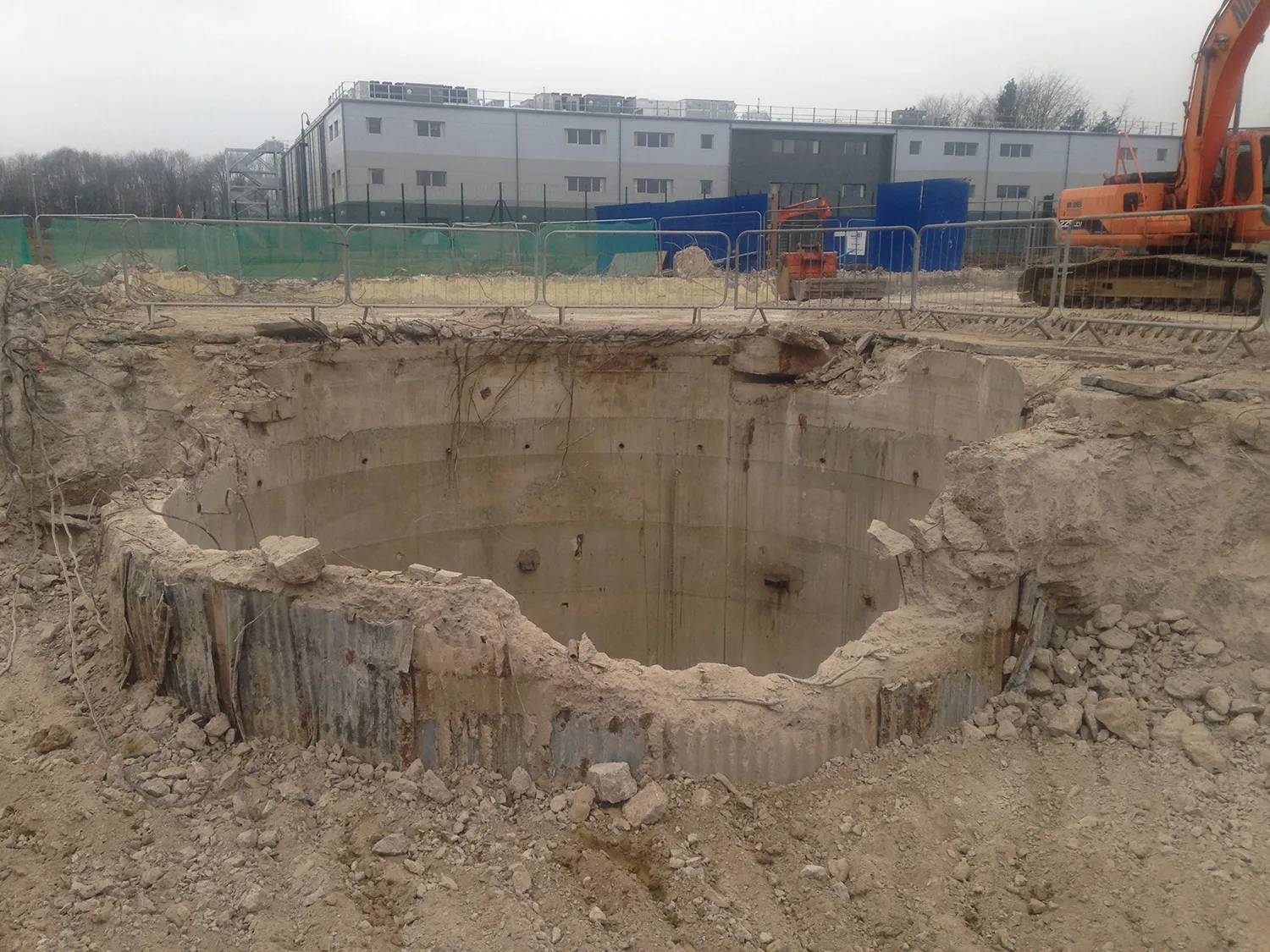 Excavated circular concrete shaft with corrugated metal lining exposed at a demolition site, excavator nearby