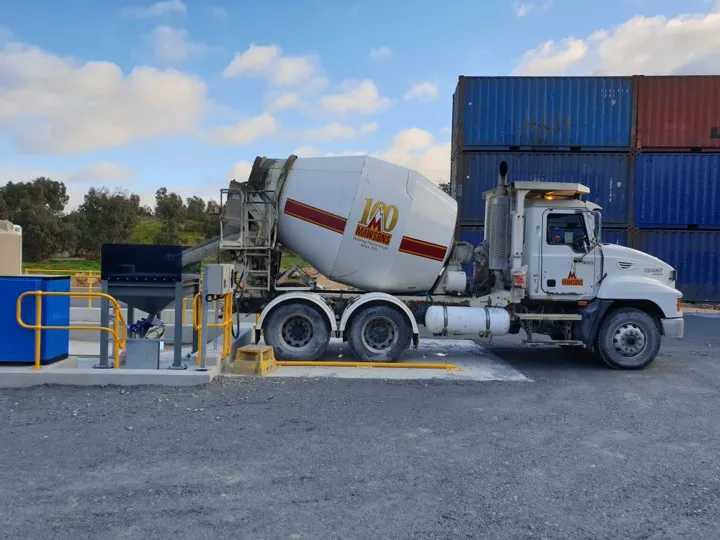 Concrete mixer truck loading at a batching plant beside stacked shipping containers on site