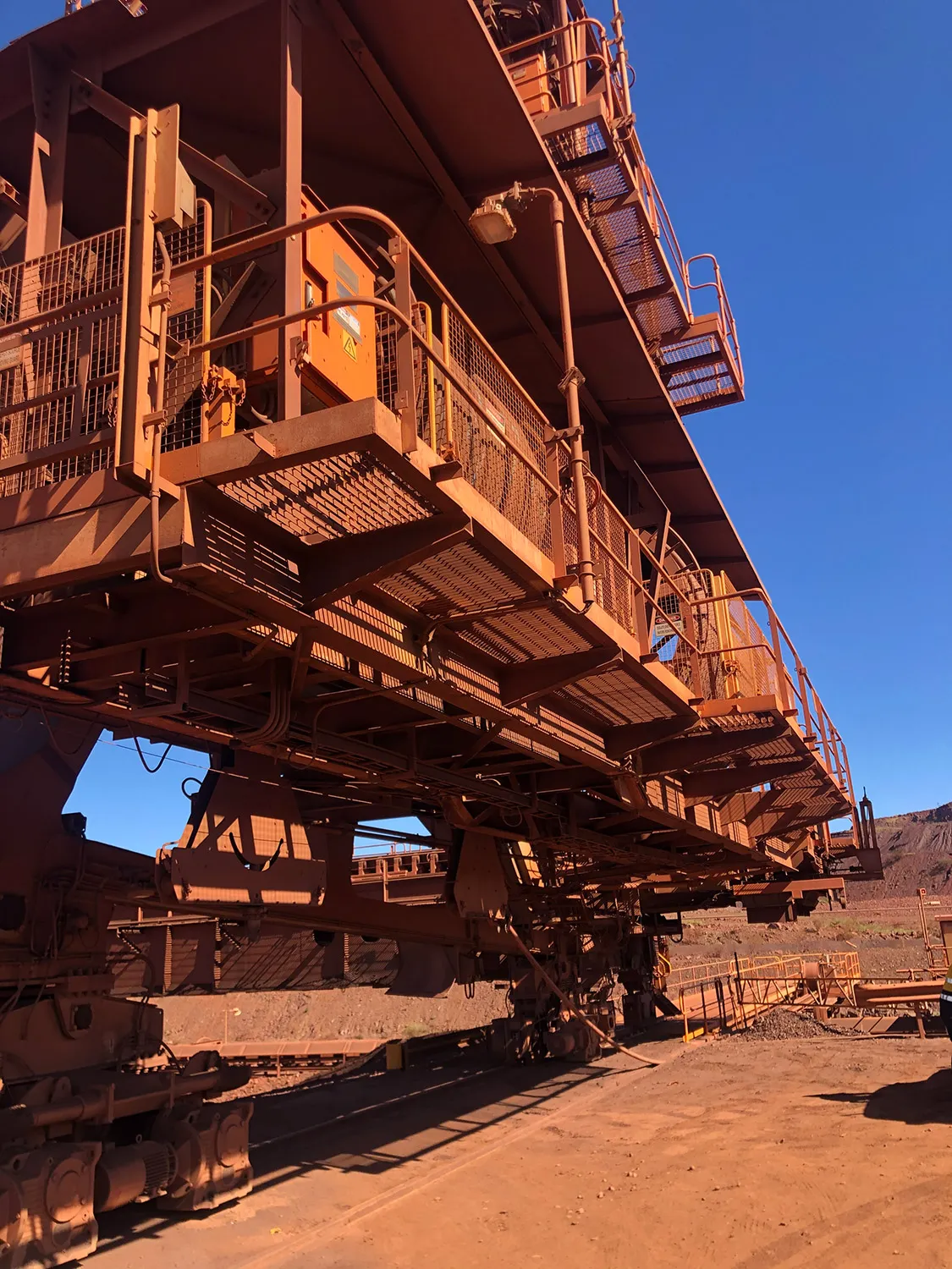 Underside view of a large orange stacker-reclaimer showing walkways and steel structure at an iron ore mine site