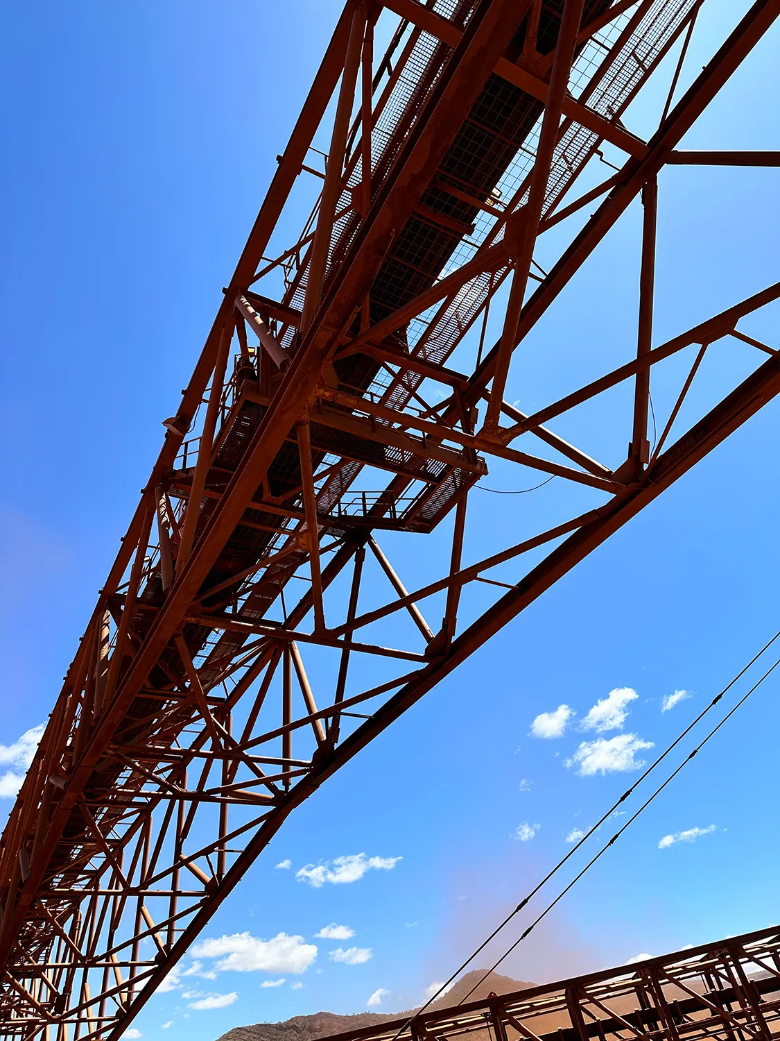Upward view of a rusted steel stacker conveyor truss and walkway against a clear blue sky